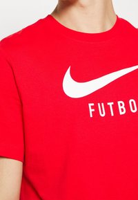 Osoba v červeném tričku s krátkým rukávem, na kterém je bílé logo Nike a slovo "FUTBOL" vytištěné bílou barvou na hrudi.