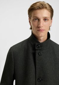 Mørkegrå uldfrakke med høj krave, subtil hørmønster og to sorte knapper foran. Minimalistisk design, skræddersyet pasform.