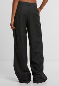Personne debout de dos face à la caméra, portant un pantalon noir ample avec une seule poche arrière, sur un fond uni clair.