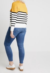 Pull rayé jaune, noir et blanc, avec un col rond. Porté avec un jean skinny bleu et des chaussures blanches à lacets avec une semelle beige.