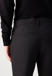 Pantalones negros ajustados con una textura suave, que cuentan con dos bolsillos traseros y un sutil detalle de costura. Combinados con una blusa negra de manga larga.