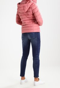 Ljusrosa dunjacka med quiltad design och huva, kombinerad med mörkblåa skinny jeans och vita sneakers. Sedd bakifrån.