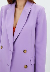 Lilat blazer, jossa on lovettu kaulus, kaksirivinen malli ja kontrastiväriset napit. Sileä kangas ja rakenteellinen siluetti.