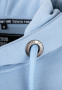 Tissu de sweat à capuche bleu clair avec cordon tressé passant à travers un œillet métallique marqué "SPITZBUB" et étiquettes de vêtement noires visibles.