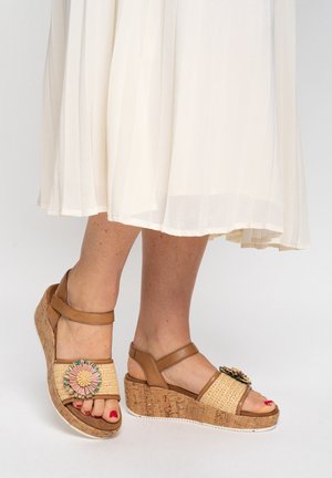 Piedi in sandali zeppa color cuoio con cinturini intrecciati e decorazione floreale, abbinati a una gonna bianca plissettata.