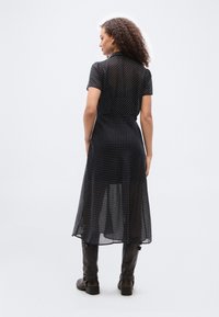 Γυναίκα με σγουρά μαλλιά που στέκεται με την πλάτη, φορώντας μαύρο φόρεμα με πουά μανίκια κοντά και μαύρες μπότες μέχρι το γόνατο.