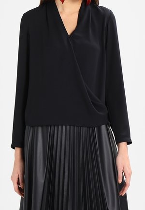 Kobieta ubrana w czarną koszulę kopertową z długim rękawem, w połączeniu z czarną plisowaną spódnicą z wysokim stanem.
