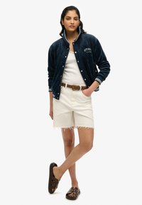 Veste bomber en velours marine avec des poignets rayés blancs, débardeur blanc, shorts effilochés beige clair et sandales à glissière brunes.
