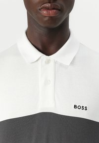 Rövid ujjú polo póló, fehér színű, szürke csíkkal. Bordás gallér és gombok. Kis "BOSS" logó fekete színben a bal mellkas területen.
