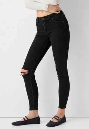 Woman wearing high-waisted black skinny jeans with distressed knee and black woven Mary Jane flats, standing with arms crossed.