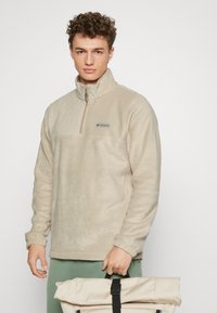Beżowy polarowy sweter z ćwierć zamkiem, z logo na piersi, ściągaczami i miękką fakturą. Model trzyma jasną torbę.