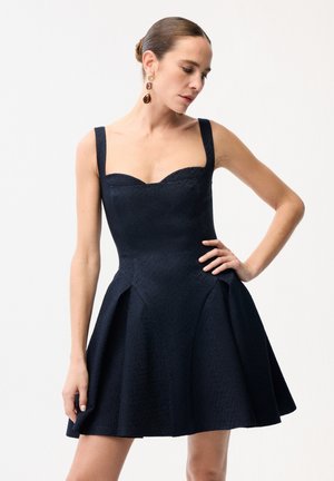 Kobieta w czarnej, teksturowanej sukience o kroju litery A bez rękawów, z dekoltem w kształcie serca, pozująca z jedną ręką na biodrze, nosząca duże wiszące kolczyki.