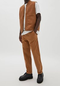 Brauner Baumwollweste mit Reißverschluss und zwei vorderen Taschen, kombiniert mit passenden braunen Hosen und schwarzen Leder-Schuhen. Einfaches Design, ohne Muster.