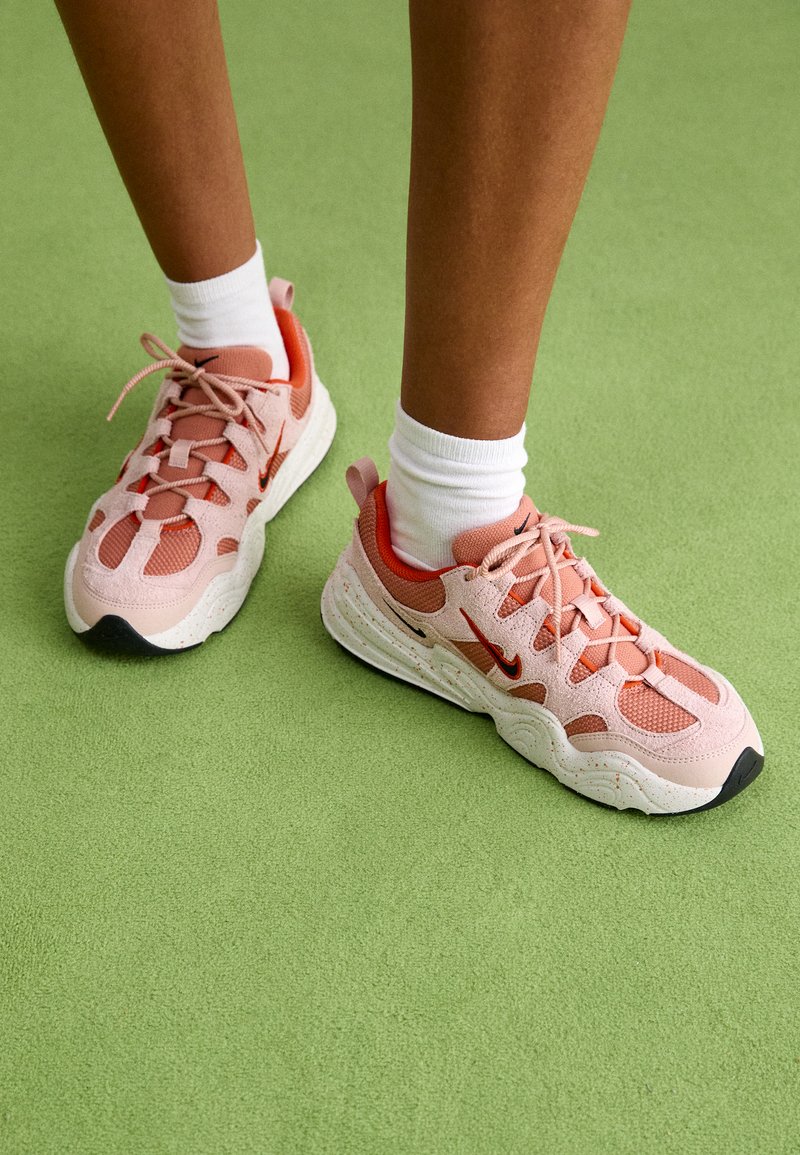 Zapatillas deportivas de color rosa claro con parte superior de malla y detalles en rojo. Tienen una suela texturizada y detalles en negro cerca de la puntera. Se llevan con calcetines blancos.