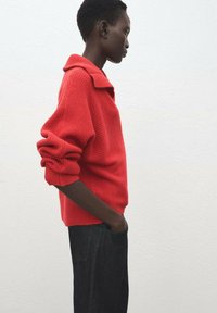 Jersey de punto rojo con cuello con solapa y puños acanalados, combinado con pantalones vaqueros oscuros. Diseño sencillo, tela texturizada y ajuste relajado.