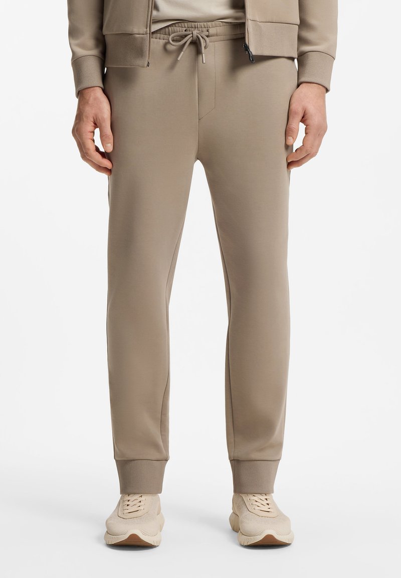 Pantalon de jogging beige avec ceinture à cordon, poignets côtelés, porté avec une veste assortie et des baskets beiges sur fond blanc.