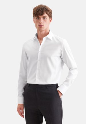 Un homme pose en chemise blanche boutonnée et pantalon foncé devant un fond uni et clair, une main dans la poche.