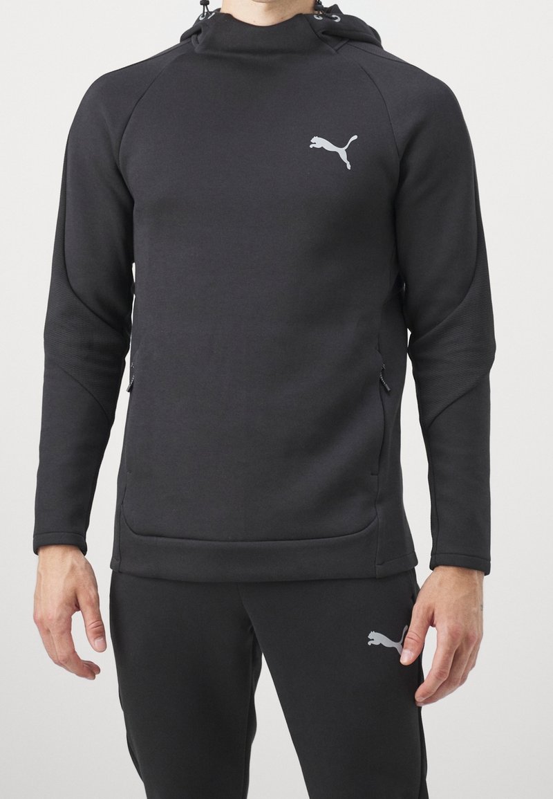 Sweat-shirt à capuche noir avec une coupe ajustée, des manches texturées, et un logo gris sur la poitrine. Dispose de poches latérales et d'un tissu lisse.