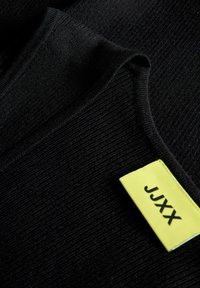 Μαύρο υφασμάτινο ριμπ με κίτρινη ορθογώνια ετικέτα που δείχνει το κείμενο "JJXX" με μαύρα γράμματα.