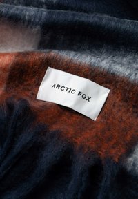 Couverture en laine mélangée à carreaux présentant des nuances de bleu marine, d'orange et de gris, avec une étiquette blanche portant l'inscription "ARCTIC FOX" cousue sur le bord.