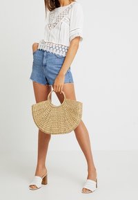Tops bianco con dettagli in pizzo, pantaloni corti in denim blu, borsa in paglia intrecciata a forma di mezzaluna e sandali bianchi con tacco a blocco.