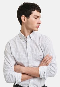 Camisa blanca de botones con un pequeño patrón azul, de mangas largas y con un bolsillo en el pecho. La tela tiene una textura suave. Cuello corto.