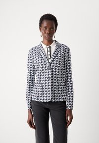 Blazer navy e bianco con motivo, design aderente, chiusura a un bottone e colletto a lancia, abbinato a una camicia bianca con colletto.