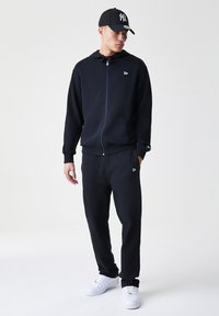 Haut de survêtement noir zippé avec cordon de serrage, associé à un jogging assorti. Des sneakers blanches et une casquette noire avec un logo complètent le look.