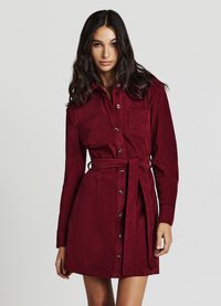 Vestito camicia in velluto a coste color bordeaux con maniche lunghe, chiusura frontale con bottoni, due tasche sul petto e vita con cintura, caratterizzato da una texture a coste.