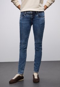 Jeans ajustados azules de denim, con un lavado intermedio, bolsillos delanteros y cierre de botón. Combinados con zapatillas de deporte marrón oscuro.