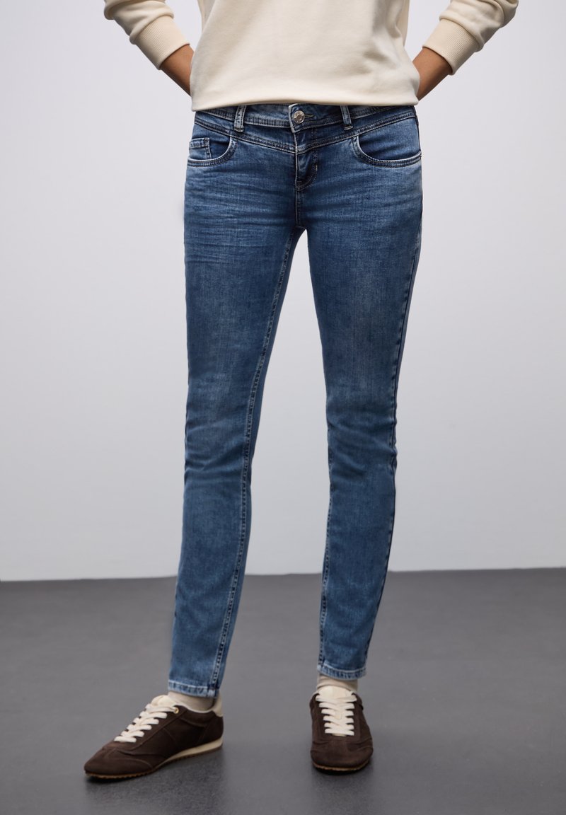 Jeans ajustados azules de denim, con un lavado intermedio, bolsillos delanteros y cierre de botón. Combinados con zapatillas de deporte marrón oscuro.