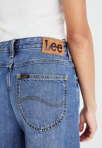 Jean en denim bleu clair avec un design classique à cinq poches, doté d'une étiquette en cuir marron avec "Lee" au dos de la taille et de surpiqûres dorées.