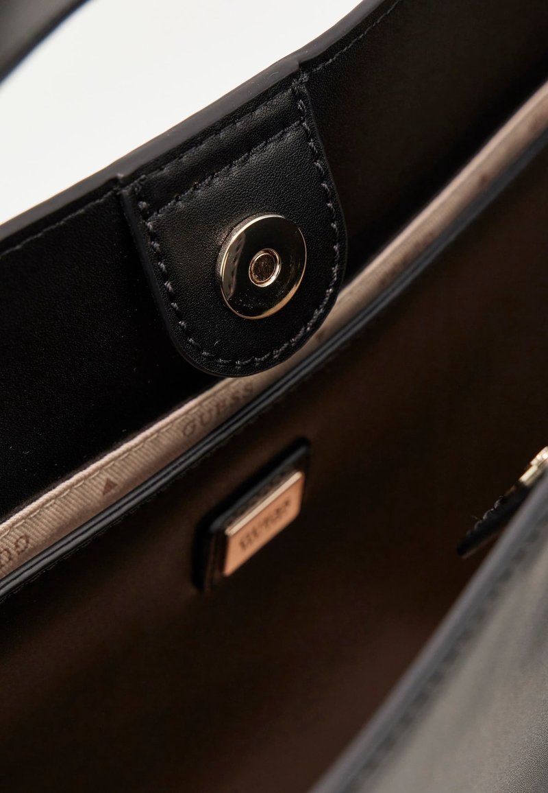 Intérieur d'un sac à main en cuir noir avec ferrures dorées, fermeture par bouton, et doublure en tissu beige ornée d'un motif de logo répétitif.