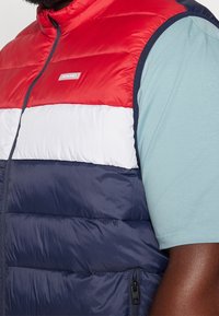 Farbenblockierte Steppweste in Rot, Weiß und Marineblau mit Reißverschluss, ausgestattet mit einem Logopatch und puffiger Textur. Darunter ein blaues T-Shirt.
