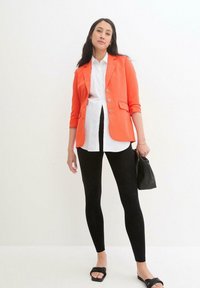 Blazer orange sur une chemise blanche, associé à des leggings noirs et des sandales noires. Le blazer a un design ajusté et deux poches à l'avant.