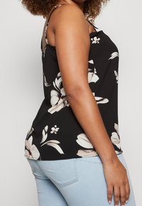 Svart ärmlös topp med stora vita blommönster, en avslappnad passform och justerbara axelremmar, i kombination med ljusblå denimjeans.