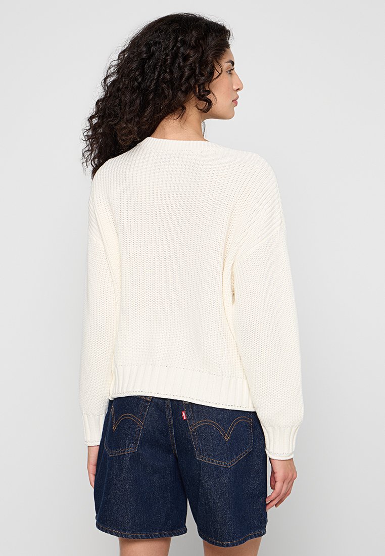 Witte gebreide sweater met een losse pasvorm, geribbelde manchetten en zoom, getoond met blauwe denimshorts met achterzakken. Gelaagd patroon door het hele stuk.
