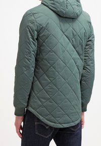 Giacca verde trapuntata con cappuccio. Presenta motivi a rombi e un orlo leggermente curvato. Il tessuto appare liscio e imbottito.