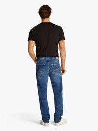 Blaue Denim-Jeans mit geradem Bein, ausgestattet mit zwei Gesäßtaschen und einem hellbraunen Lederpatch am Bund. Kombiniert mit einem schwarzen T-Shirt.