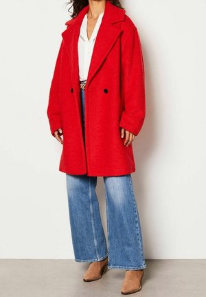 Donna che indossa un cappotto in lana oversize di un rosso acceso, una camicetta bianca, jeans blu a gamba larga e stivaletti in camoscio color nocciola, in piedi.