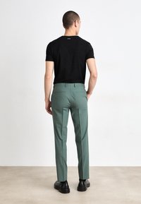 Zwarte overhemd met korte mouwen, groene op maat gemaakte broek met een gladde textuur, twee achterzakken en zwarte schoenen. Neutrale achtergrond.