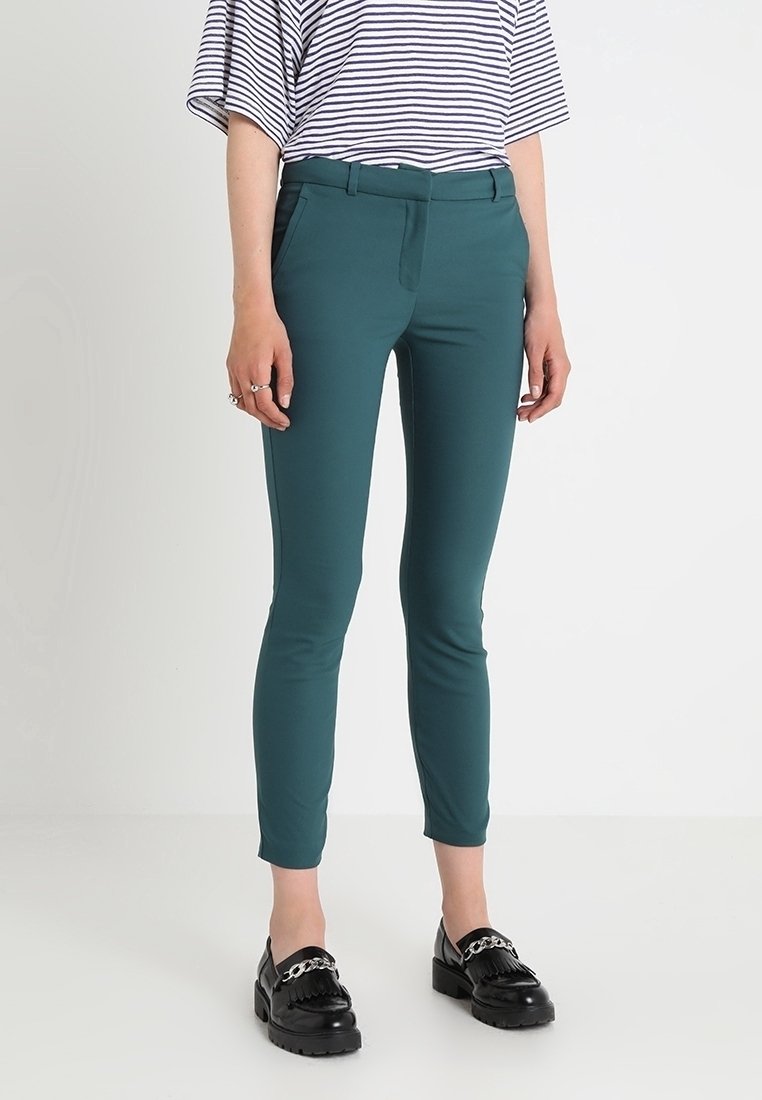 Personne portant un pantalon slim sarcelle aux chevilles, des mocassins noirs avec détail chaîne, et un top rayé blanc et bleu marine à manches courtes.