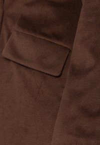 Brunt stof med en struktureret overflade, der har en rektangulær lomme. Materialet virker blødt og varmt med et subtilt mønster.