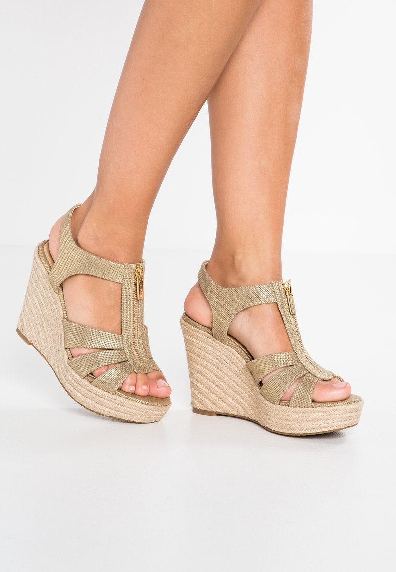 Femme portant des sandales compensées tissées beige avec une fermeture éclair à l'avant et plusieurs brides, debout sur une surface blanche.