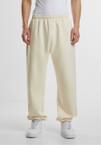 Persona que lleva joggers color crema con puños y cinturilla elásticos, zapatillas blancas y una camiseta sin mangas blanca contra un fondo liso.