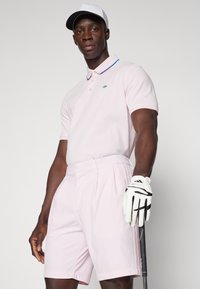 Camisa polo rosa claro com uma risca azul na gola, combinada com calções de golfe a condizer. Luvas brancas e um taco de golfe completam o conjunto.
