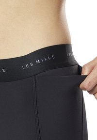 Czarne legginsy sportowe z szerokim elastycznym pasem, na którym widnieje logotyp "LES MILLS". Materiał wydaje się gładki i elastyczny, dobrze dopasowując się do sylwetki.