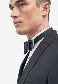 Schwarze Smokingjacke mit Satinrevers, weißes Hemd mit Fliege, zeigt eine maßgeschneiderte Passform und klare Linien. Profilansicht eines männlichen Models.