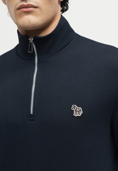 Άνδρας που φοράει σκούπερο μπλε σκούρο με φερμουάρ μέχρι το στήθος και πολύχρωμο ριγέ λογότυπο αλόγου κεντημένο στο αριστερό στήθος.