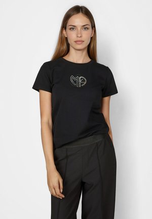 Junge Frau mit langen hellbraunen Haaren trägt ein schwarzes T-Shirt mit kurzen Ärmeln und silbernem "MA"-Logo sowie schwarze Hosen, steht vor einem weißen Hintergrund.